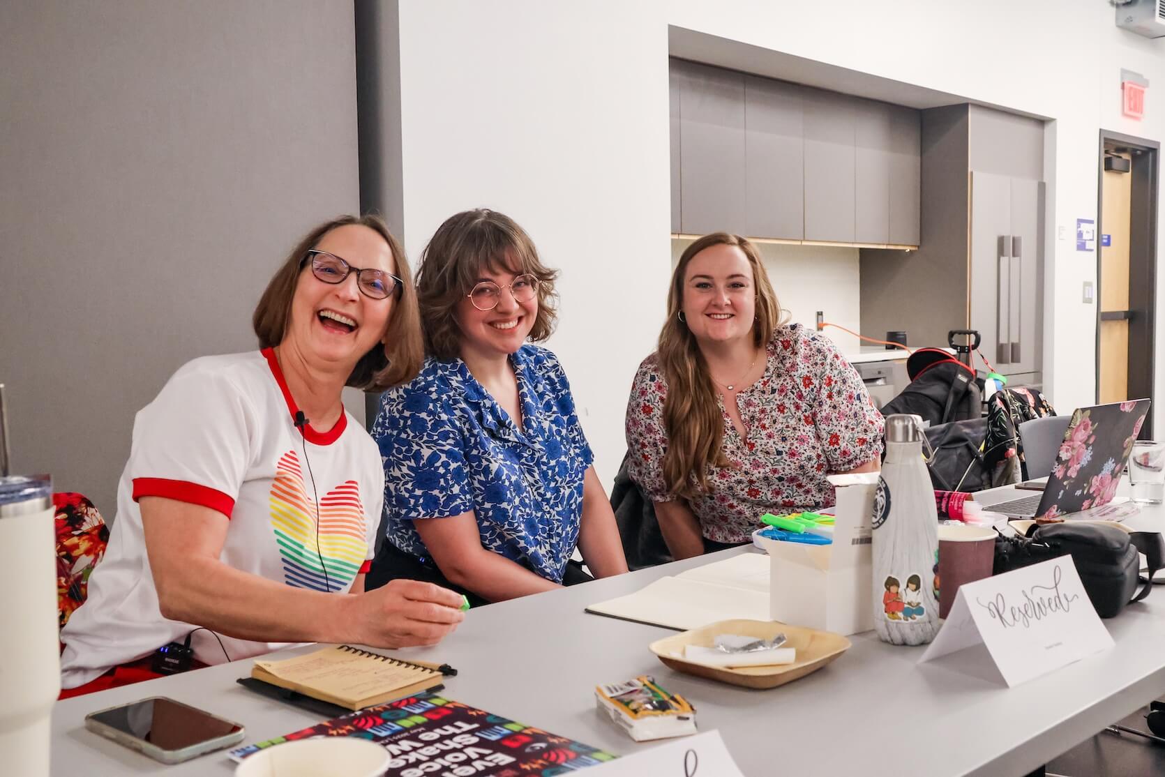 Three white women sit together at a table, smiling. On the left is an older woman with bob length brown hair. She is wearing glasses and a t-shirt with a rainbow heart on it. She is smiling wide. The woman in the middle has short curly bob length hair with bangs. She is also wearing glasses, and a white and blue floral button up blouse. The woman on the right has long wavy reddish-blonde hair, and she is wearing a floral blouse. Laptops and notebooks and other materials are laid out on the table.