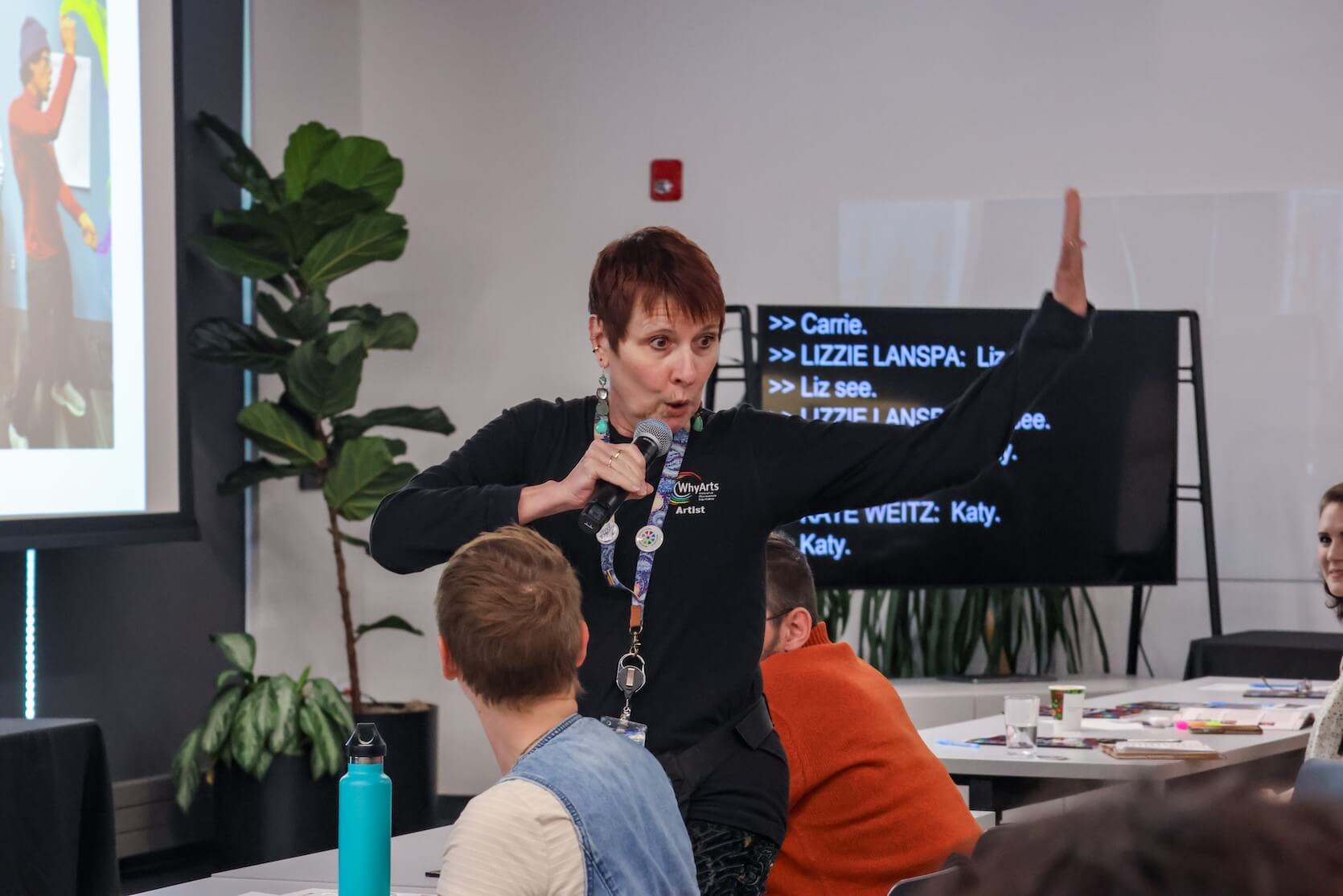 A white woman with short pixie red hair, wearing all black. She is holding a microphone and in the middle of moving around as she speaks to the audience in the conference room.