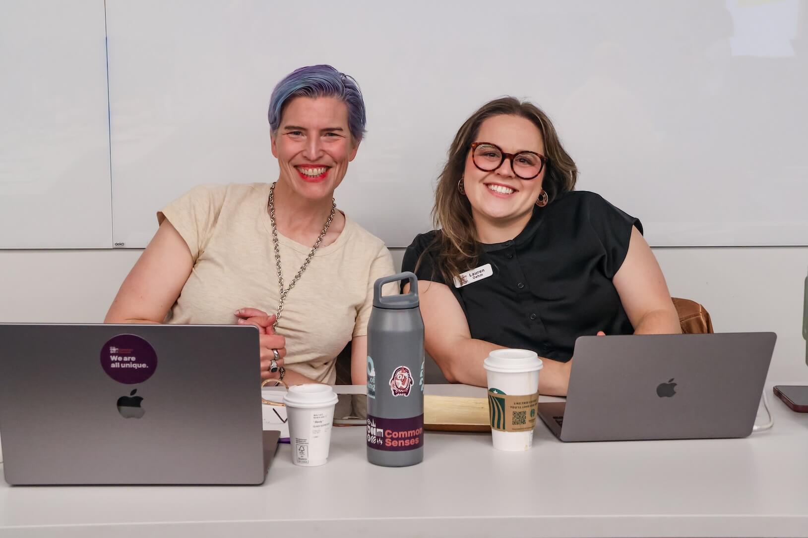 Two white women sitting together at a table, smiling. The woman on the left has short blue and purple hair, wearing a cream t-shirt with jewelry. The woman on the right has long brown hair, wearing glasses and a black button up blouse. Their laptops are open in front of them, sitting beside a water bottle and two coffee cups.