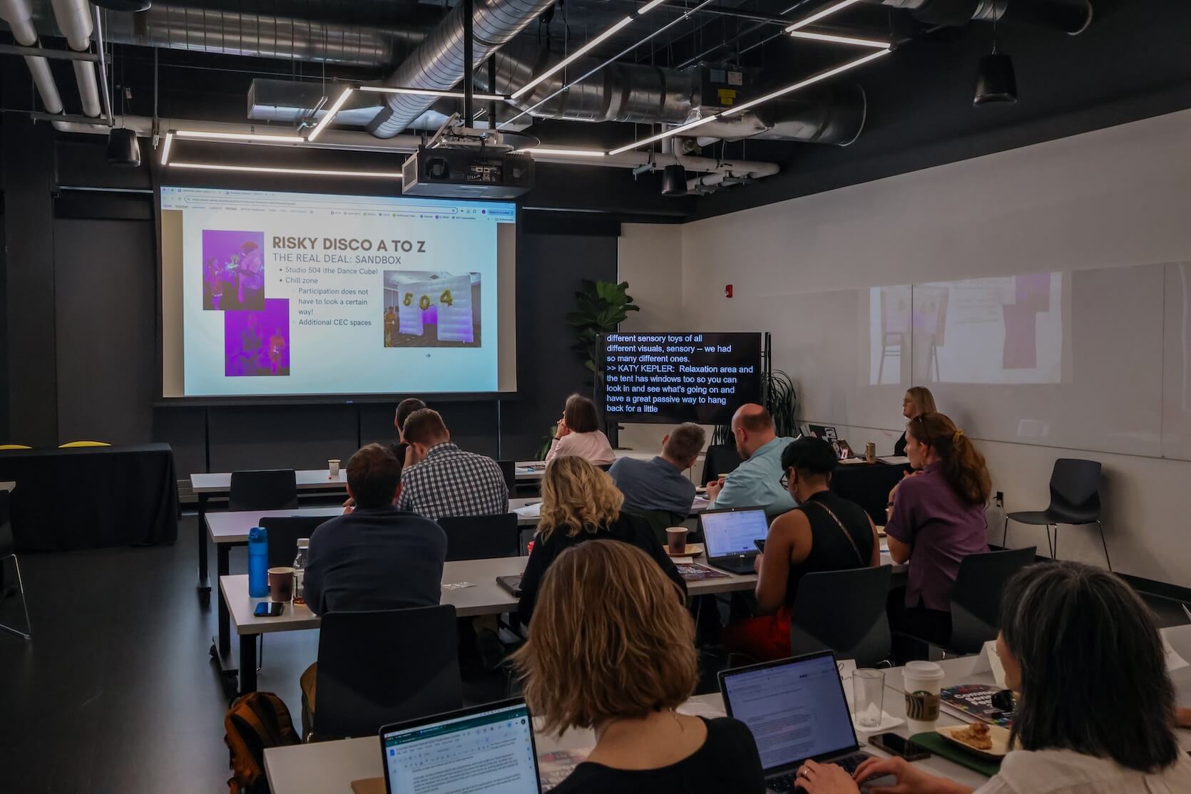 A group of people sitting in a conference room, laptops open. They are learning about Risky Disco, where a PowerPoint is showing information. Live Captions are displayed at the front of the room.