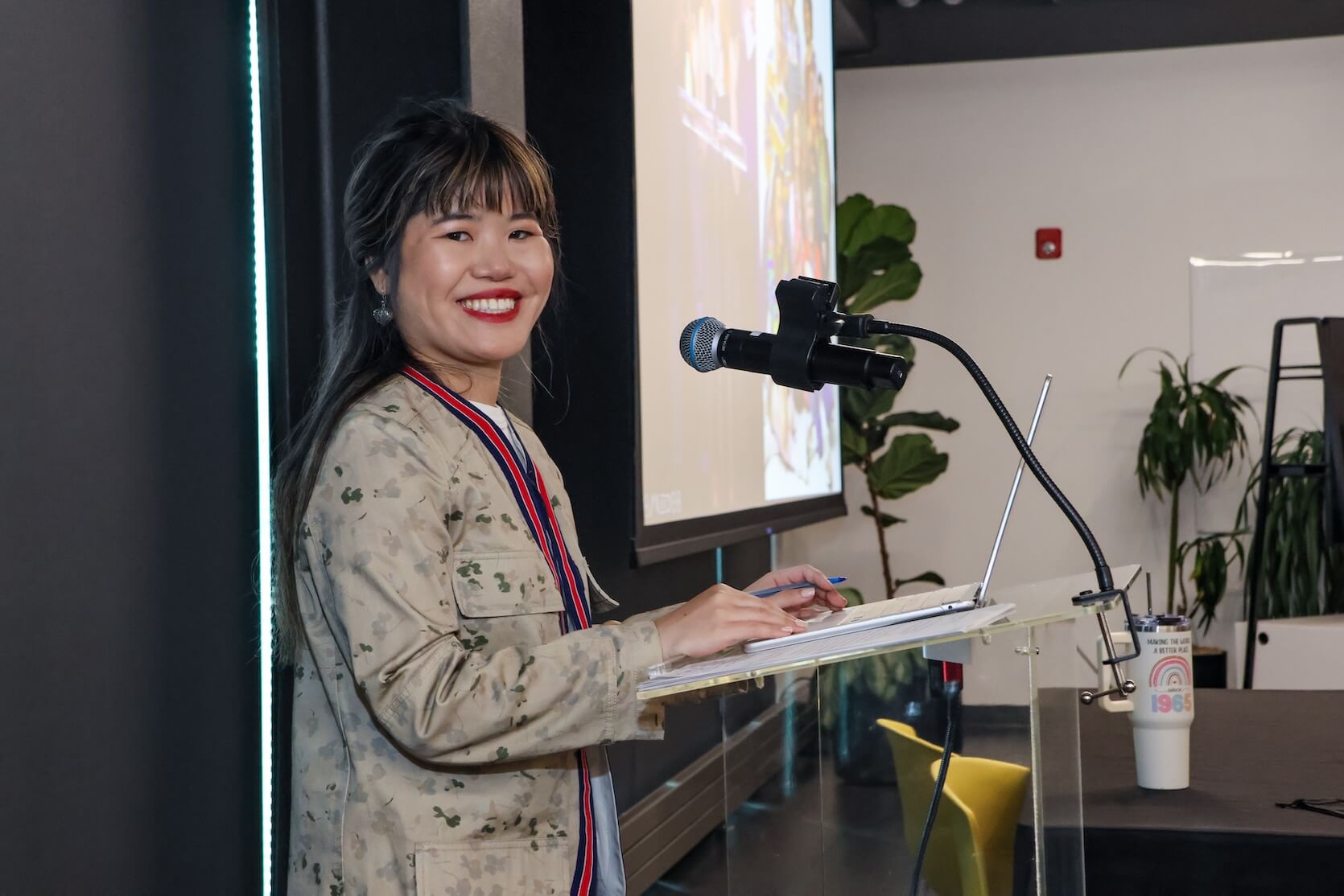 Young Asian woman with long black hair with brown bangs, half pulled back. She is wearing red lipstick, and a light camo-like long sleeve over a white shirt. She is standing at the podium, smiling at the camera. A laptop is open.