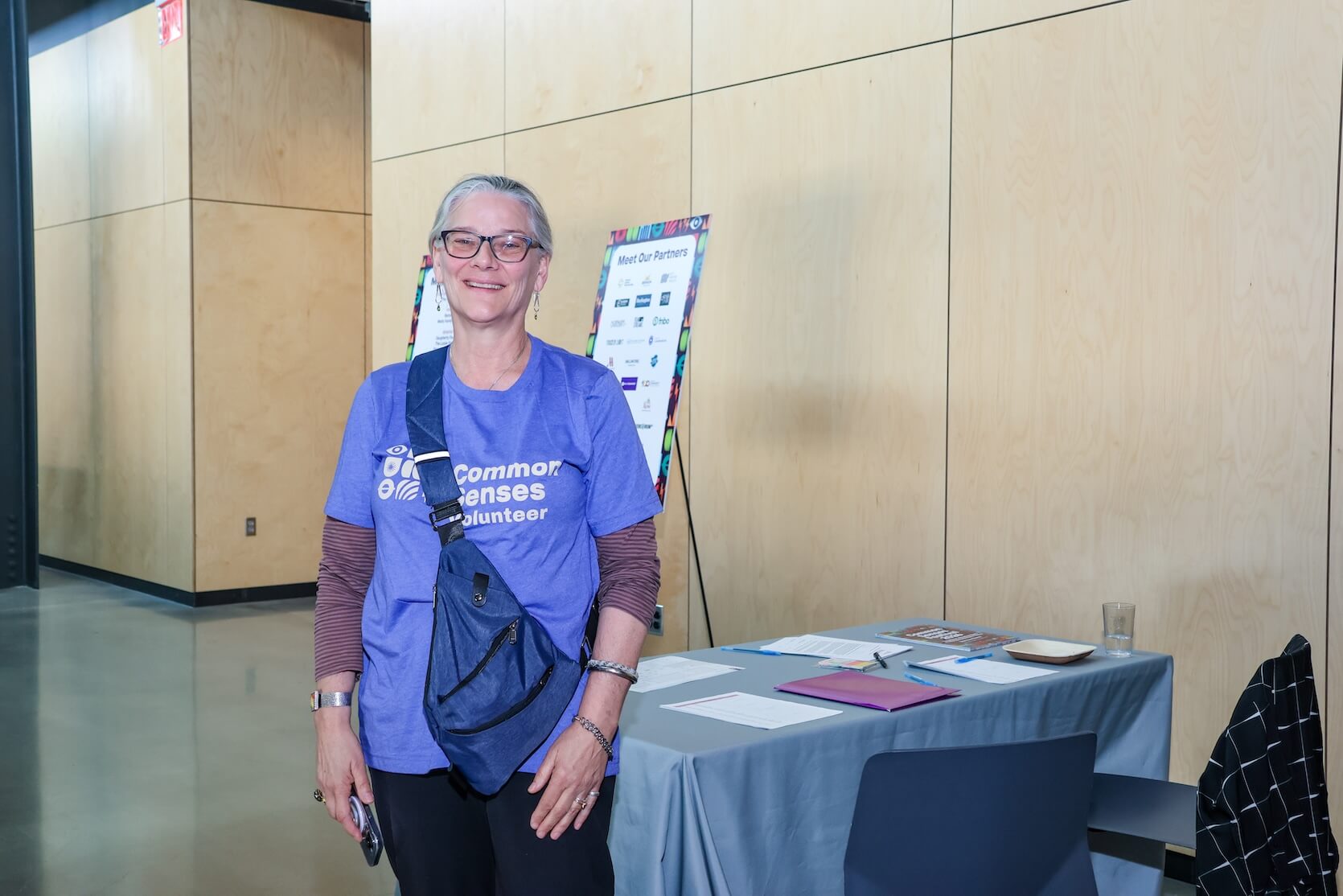 An older white woman with gray hair pulled back into a ponytail, wearing a blue Common Senses Festival volunteer t-shirt, black pants, and a black slingbag smiles at the camera. A table with event materials and sponsors posterboard are behind her.