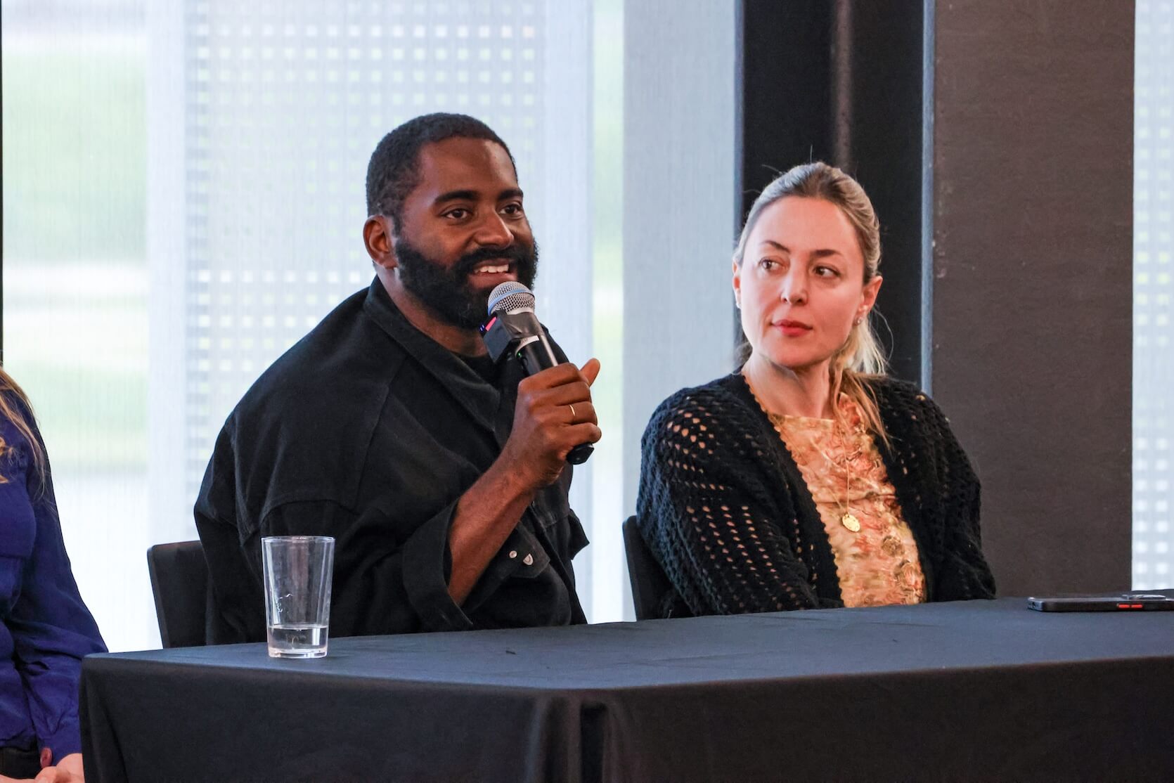 A Black man with short black hair and a beard holding a microphone, speaking. A white man with her hair pulled back sits beside him, looking at him while he speaks.