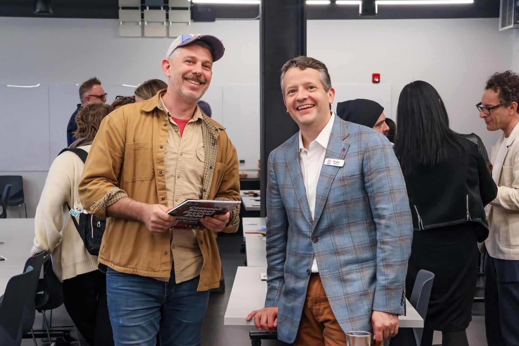 Two white men standing together, smiling. The man on the left is in casual attire, wearing a hat, various denim button ups and jeans. The man on the left is in more business attire, wearing a suit and jeans.