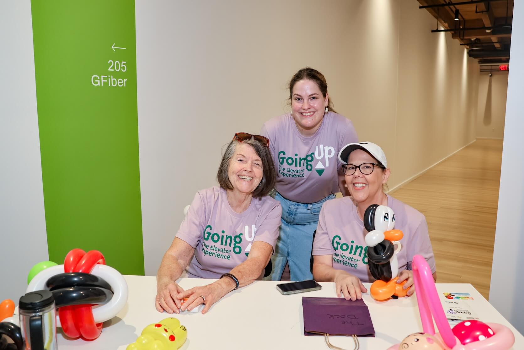 Three white women, two standing and one sitting, posed together, smiling. They are all wearing lilac Going Up The Elevator Experience t-shirts. Animal balloons are laid out onto the table.