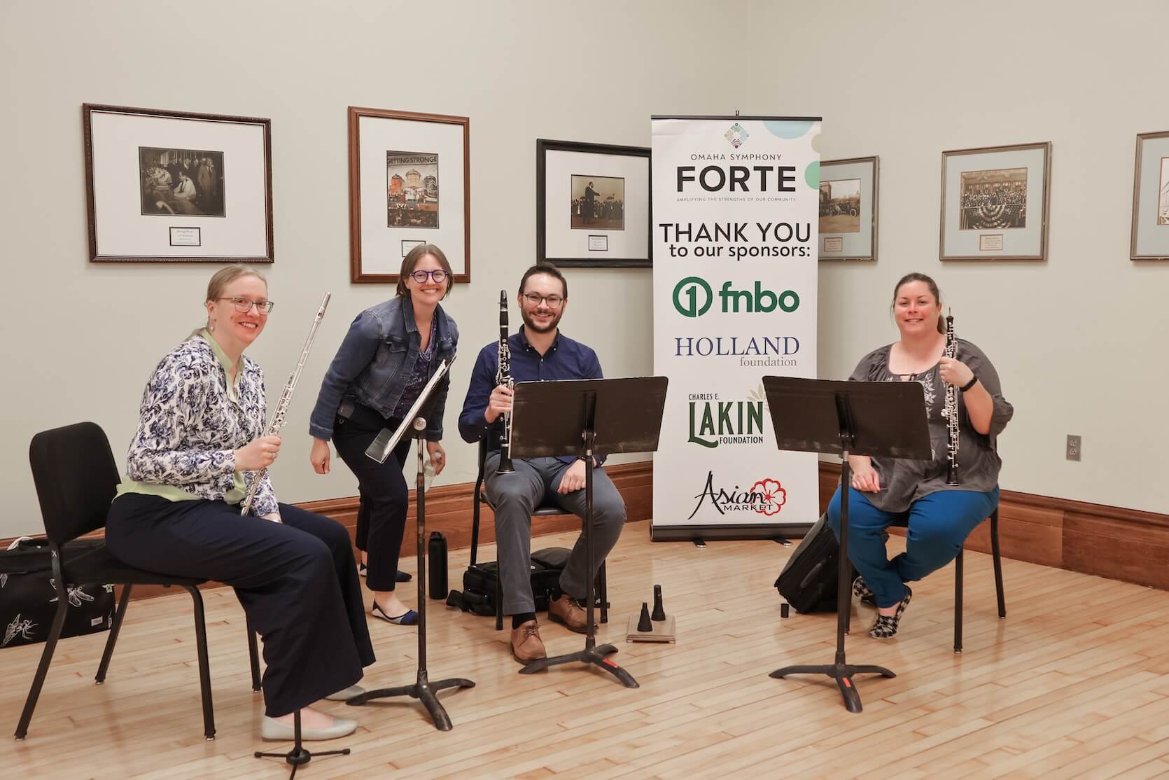 Three white women and one white man together. Two women and one man are sitting, holding their instruments. Another woman is standing, hunched over. All are smiling. Poster of sponsors and picture framed wall art are behind them.