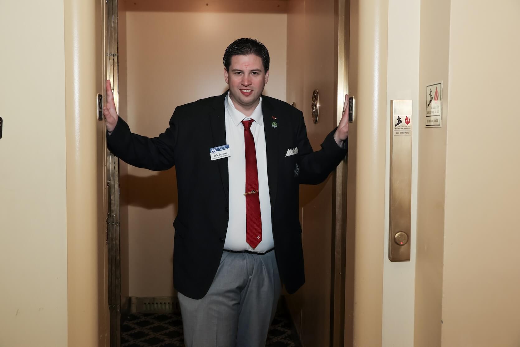 A young adult white man with black hair combed to both sides, wearing a suit, looks toward the camera while he holds the elevator door open.