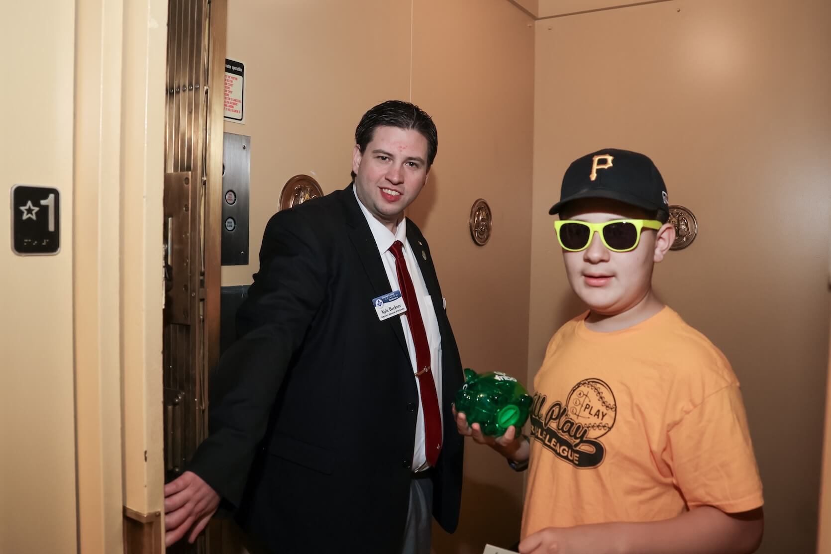 A young adult white man with black hair combed to both sides, wearing a suit, looks toward the camera while he holds the elevator door open for a young boy wearing a hat, sunglasses, and an orange graphic shirt. He is holding a small glass/plastic green pig.