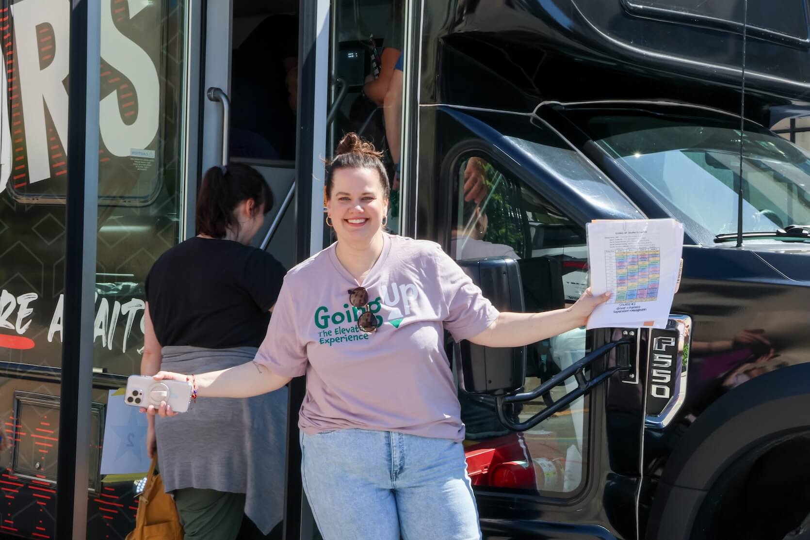 A white woman wearing a lilac Going Up The Elevator Experience t-shirt, light denim jeans, glasses on the shirt. Her hair is pulled up into a bun. She is holding some papers, arms stretched out, smiling. She is standing in front of a bus, where one person is in the process of stepping into.