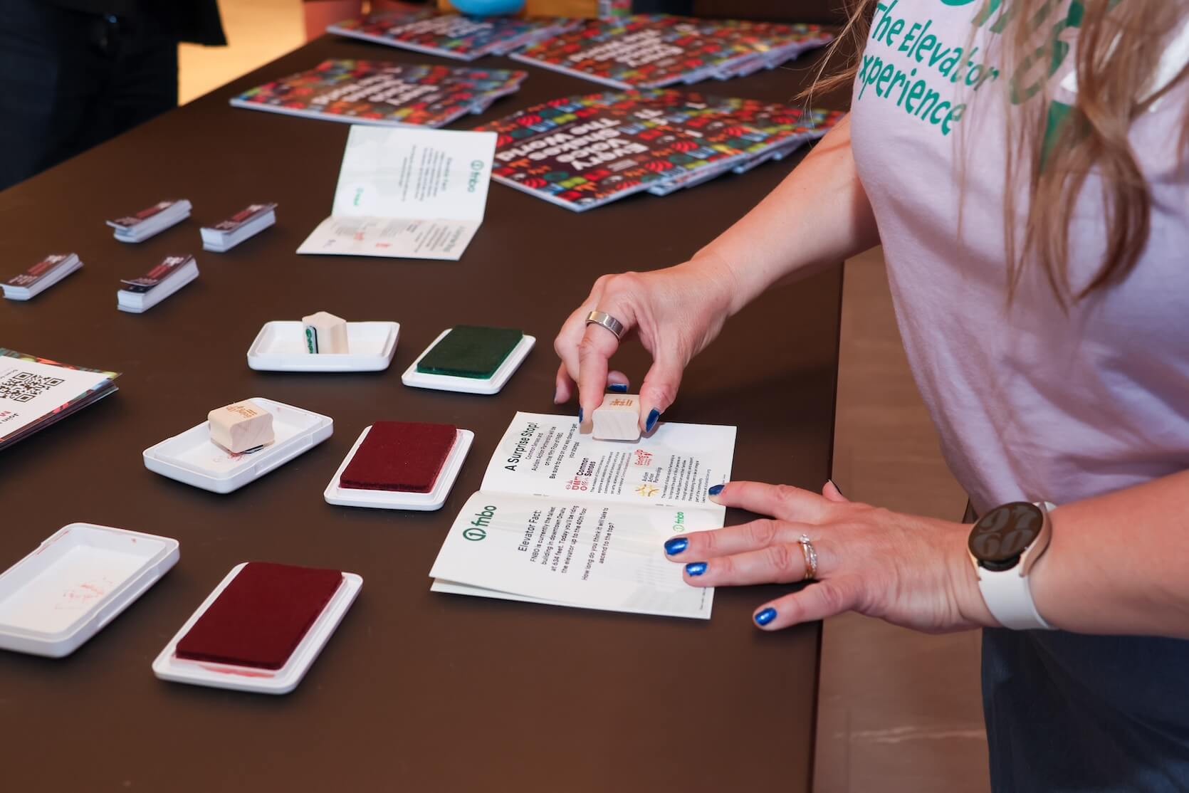 A woman is stamping something into the Going Up The Elevator Experience booklets. Lots of colored ink pads and other event materials are laid out on the table.