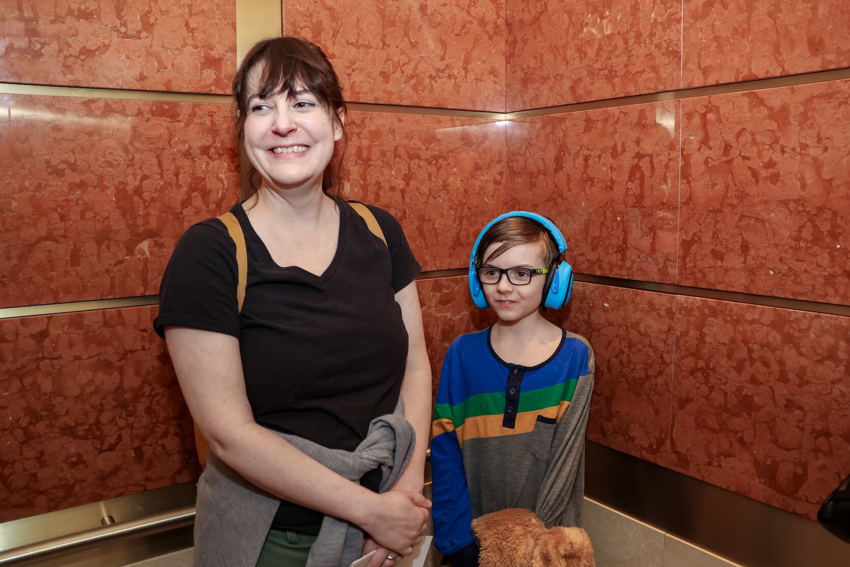 A white woman and child stand together in the elevator. The woman has brown hair pulled up into a ponytail and is wearing a black v-neck shirt and green pants with a gray shirt wrapped around her waist. She is carrying a backpack. The child is wearing headphones, glasses, and a multicolored long sleeve shirt. He looks to be holding a stuffed animal.
