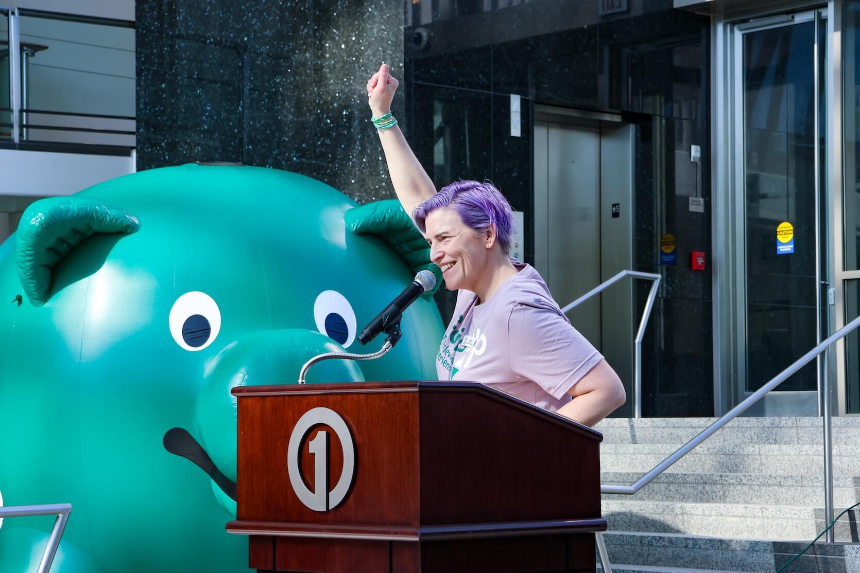 A white woman with short purple hair and a lilac Going Up The Elevator Experience t-shirt stands at the podium, smiling with her arm raised. A large blown up green pig is behind her.