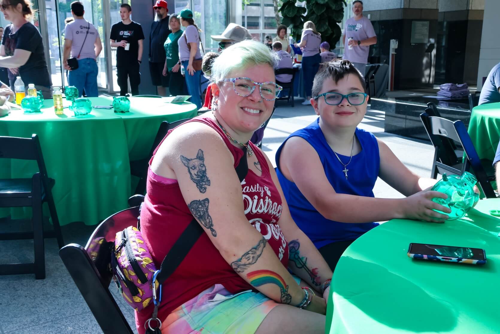 A woman and child sit at the table, smiling at the camera. The woman is wearing glasses, a red graphic tank top with pastel multicolored shorts, and has tattoos. The child is wearing glasses, a blue tank top, black shorts, and a necklace.