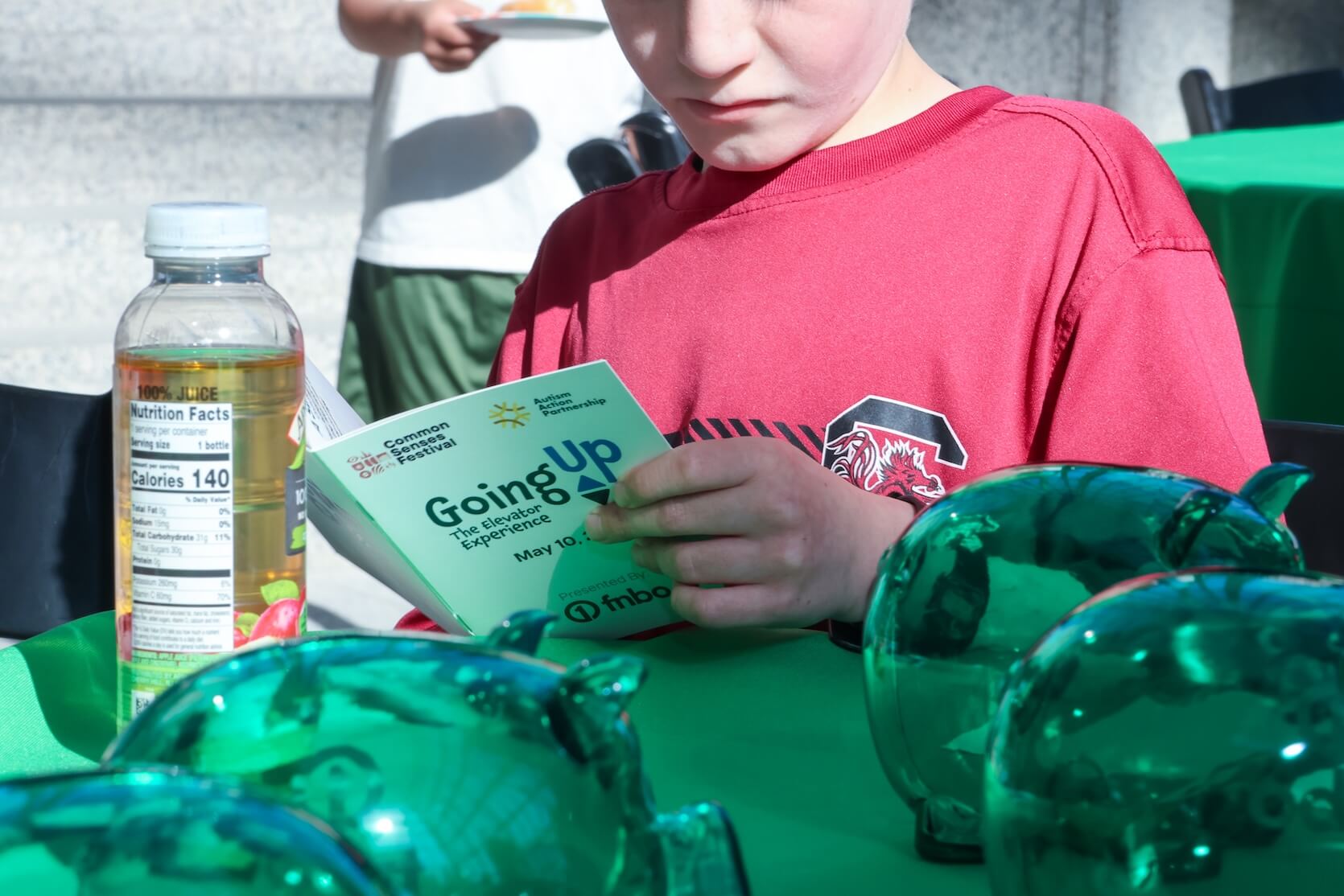 A young boy is sitting at the table reading the Going Up The Elevator Experience booklet. A bottle of apple juice and many glass/plastic pig objects are laid out on the table.