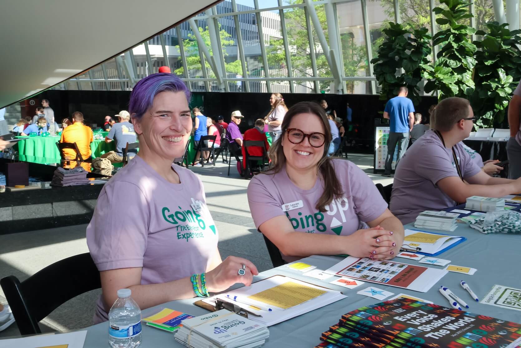 Two white women sitting together at a table, smiling. They are both wearing lilac Going Up The Elevator Experience t-shirts. The woman on the left has short purple hair. The woman on the right has long brown hair pulled half back with a clip. She is also wearing glasses. Event materials are laid out on the table. Many people are in the background.