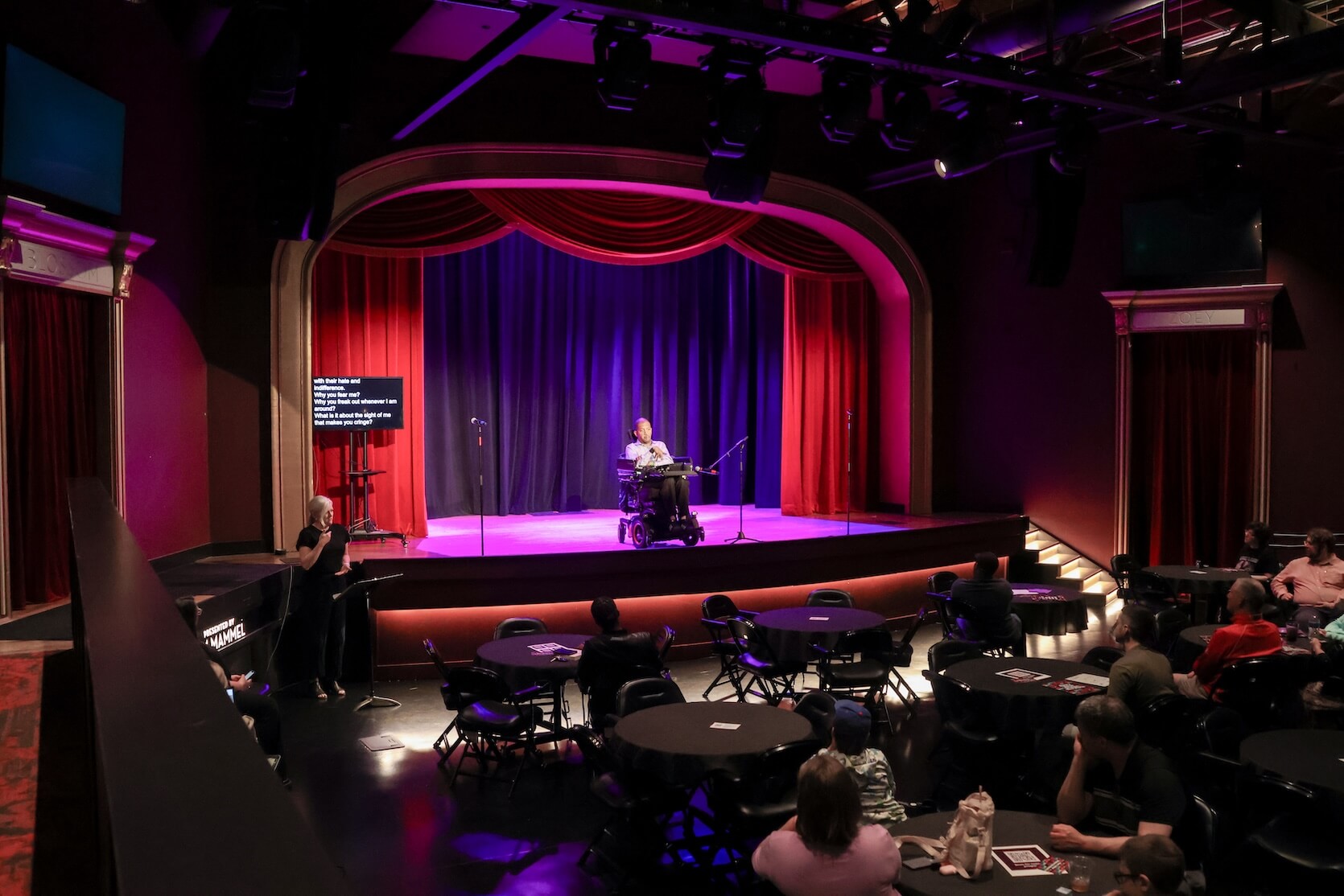 A Black man wearing a blue button up and black slacks, sitting in a powerchair on stage. Live Captions are on stage beside him, and an ASL interpreter is standing on the floor. The audience is watching him.