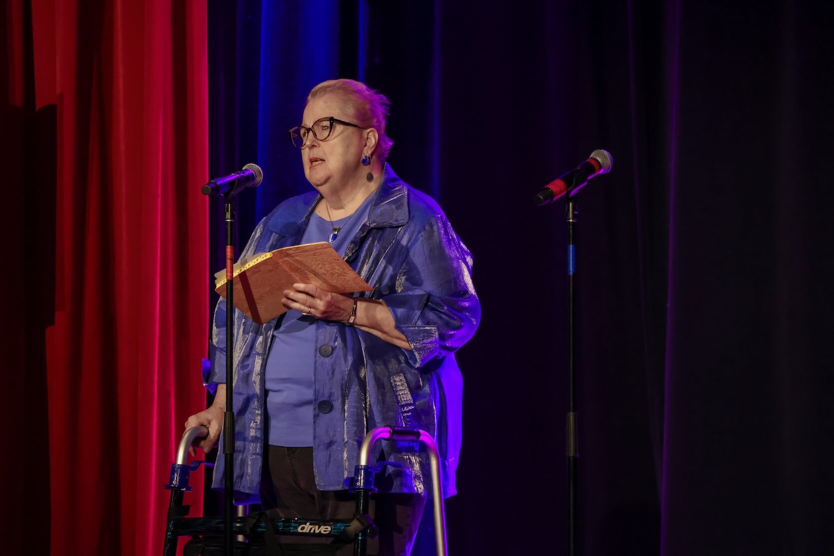 A white woman with short hair pulled back. She is wearing glasses, jewelry, a blue shirt and short sleeved button up, and black jeans. She is standing at the microphone with her walker, notebook in hand.