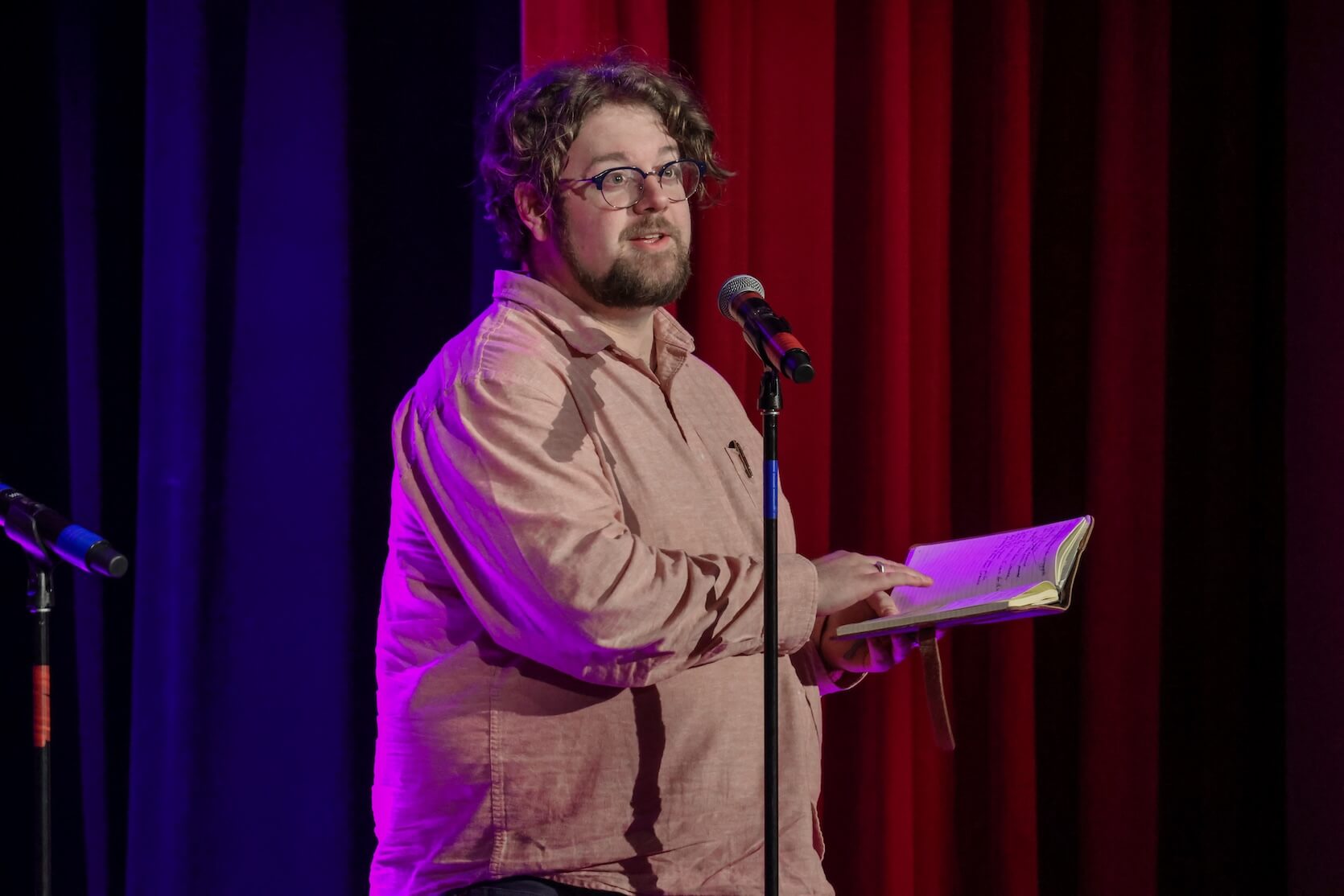 A white man with short curly brown hair and beard, wearing glasses and a light brown button up. He is holding a notebook while speaking at the microphone.