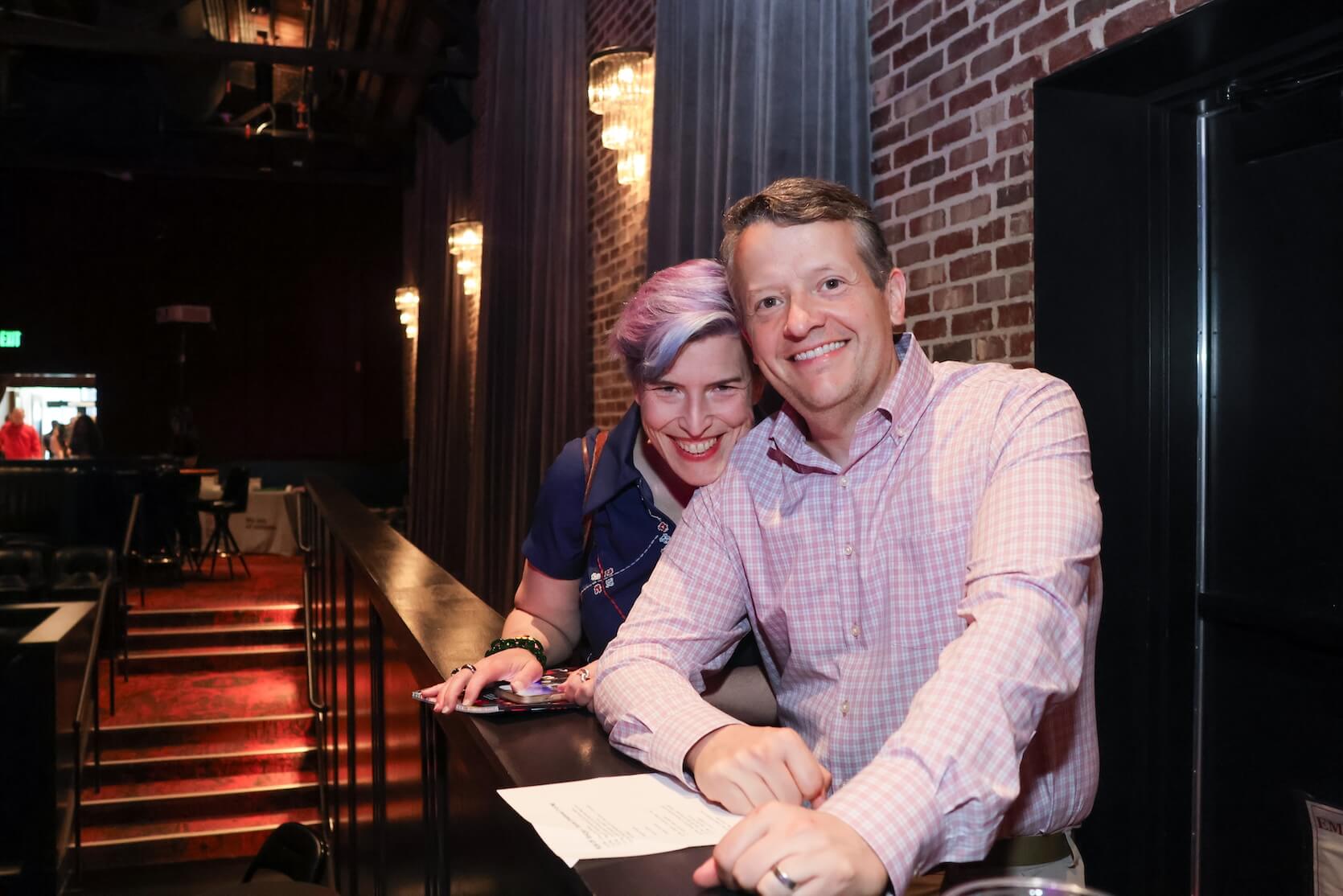 A white woman and white man standing together, smiling. The woman has short purple hair, wearing a dark blue dress. The man has short brown hair and is wearing a pink and white gingham button up. The woman is resting her chin on the man’s shoulder.