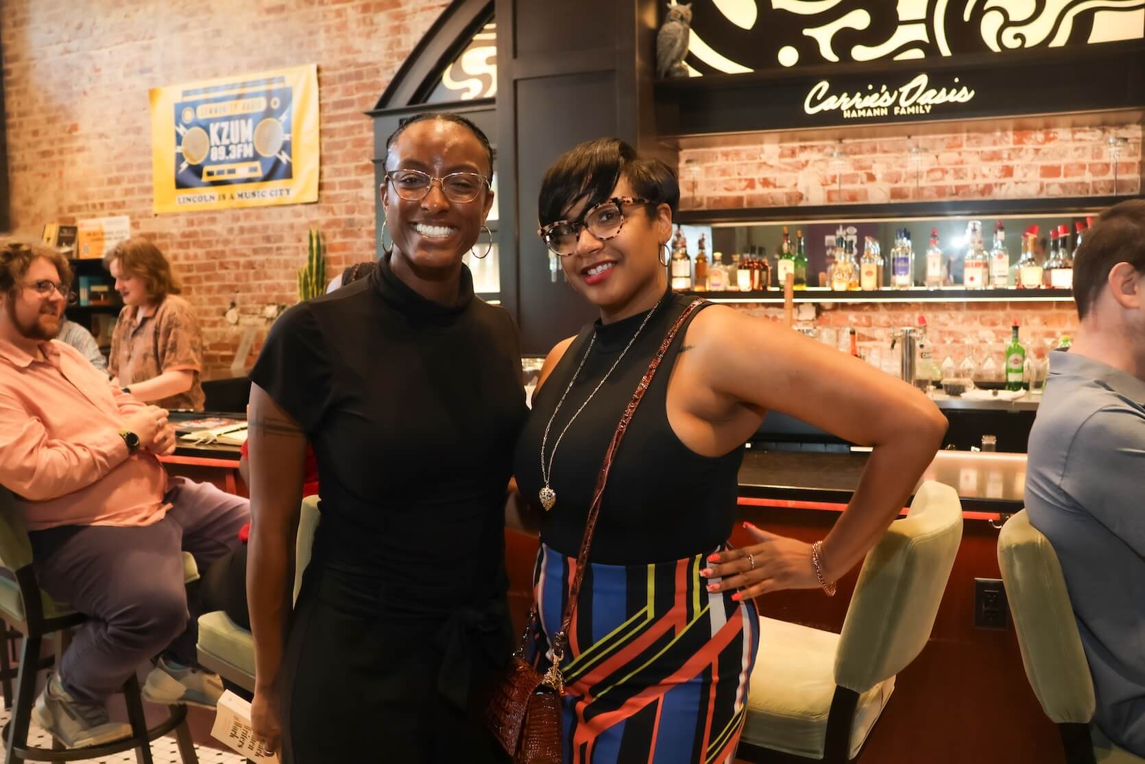 Two Black women stand together, smiling. The woman on the left has hair pulled back and is wearing a black dress. The woman on the right has short pixie hair, wearing glasses and a black tank top with a multicolored skirt.