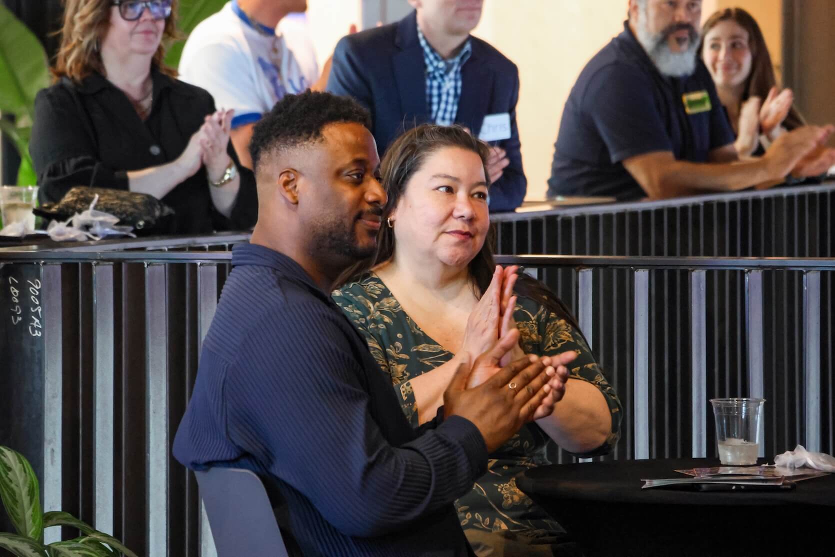 A man and woman sitting together at a table, clapping for the speaker.
