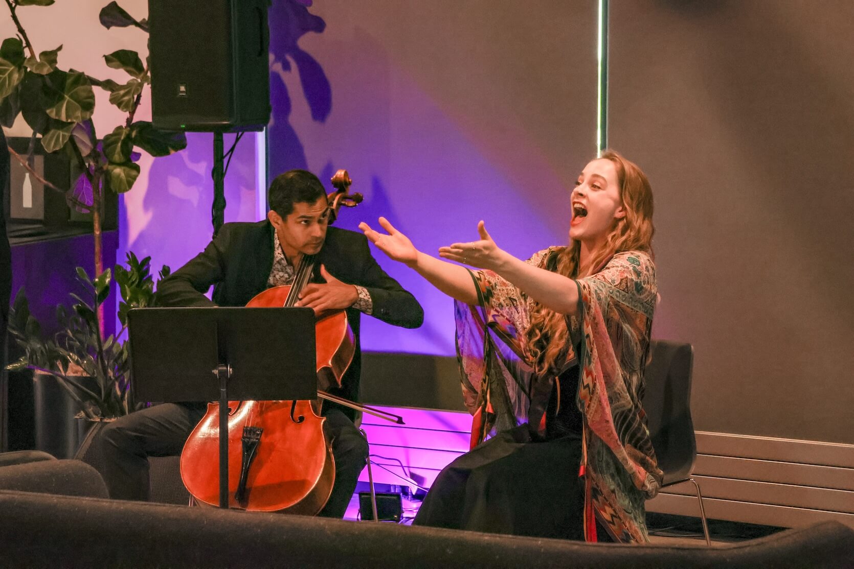 A male cellist is playing, looking at a woman who is singing, arms stretched out in front of her.