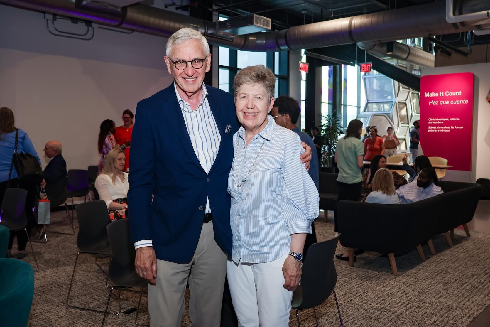 A white man and white woman stand together, one arm hug, smiling. The man is tall, with short white hair, wearing a blue and white striped shirt, blue blazer, and grey slacks. He also has glasses on. The woman has short reddish hair, wearing a blue and white striped shirt, and white slacks. More people are in the background.