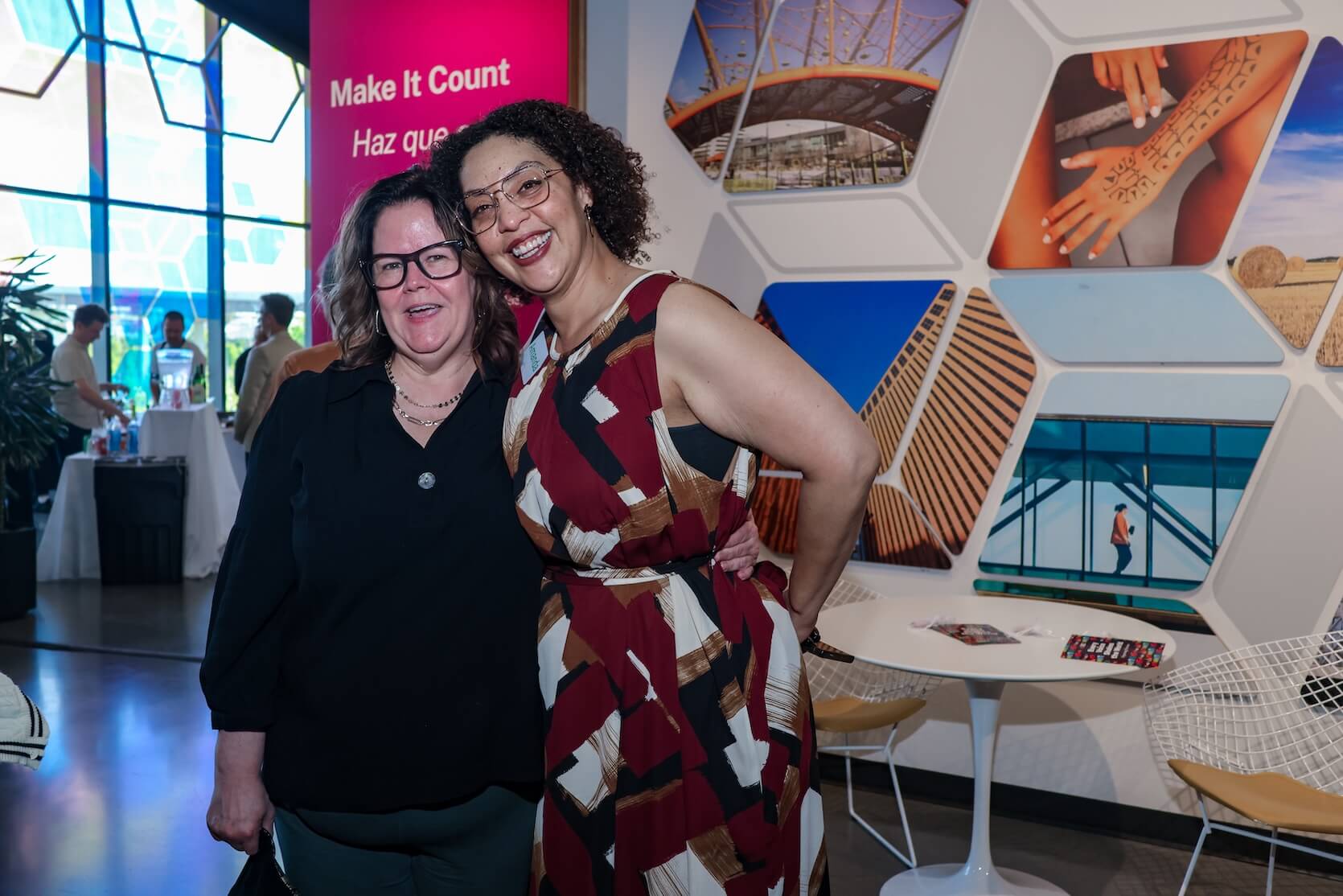 A white woman and Black woman stand closely together, one arm hug, smiling. The white woman has long curled brown hair, wearing glasses, jewelry, black shirt, and emerald banks. The Black woman has curly bob length black hair, jewelry, and a multicolored dress.