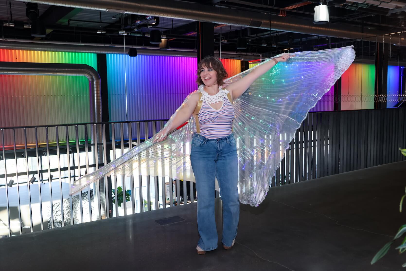 Young white woman with bob length brown hair, wearing a multicolored striped tank top with blue jeans, wearing a piece of large fabric to act as wings. Her arms are open to act as if she’s flying.