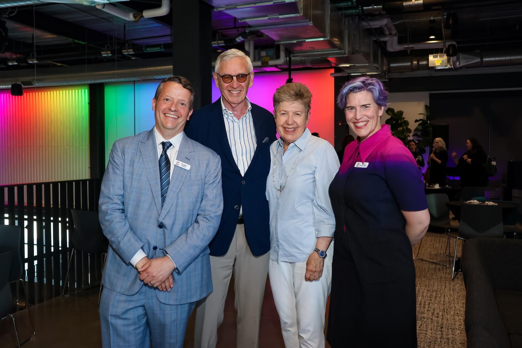 Two white men and two white men stand together, smiling at the camera. The men are wearing blue suits and have short hair: one brown, one white. One woman with short reddish hair wearing a blue and white striped button up with white pants. The woman on the far right has short purple hair, wearing a mostly black dress with small sections of pink and purple at the top.