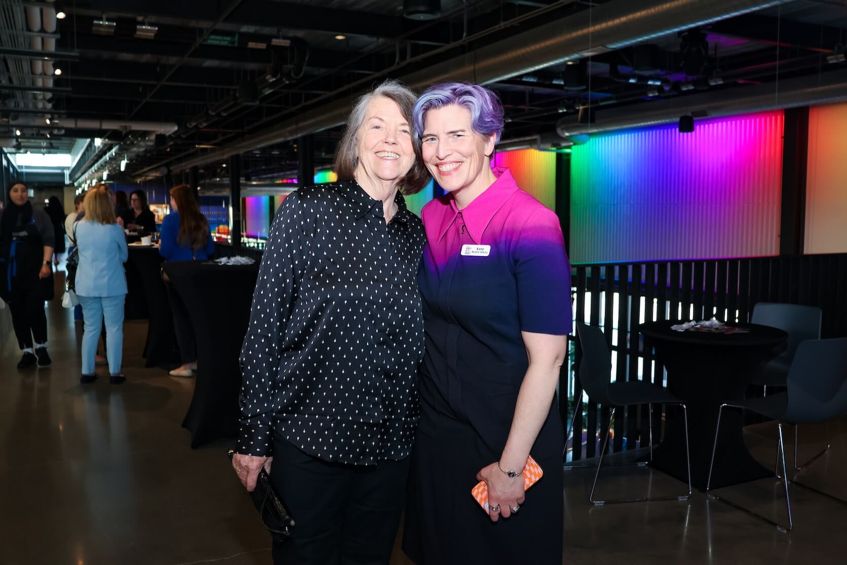 Two white women standing close together, smiling at the camera. Left has bob length brown and gray hair, wearing a long sleeve black button up with some white detail, and black pants. Right has short purple hair, wearing a mostly black dress with small sections of pink and purple at the top.