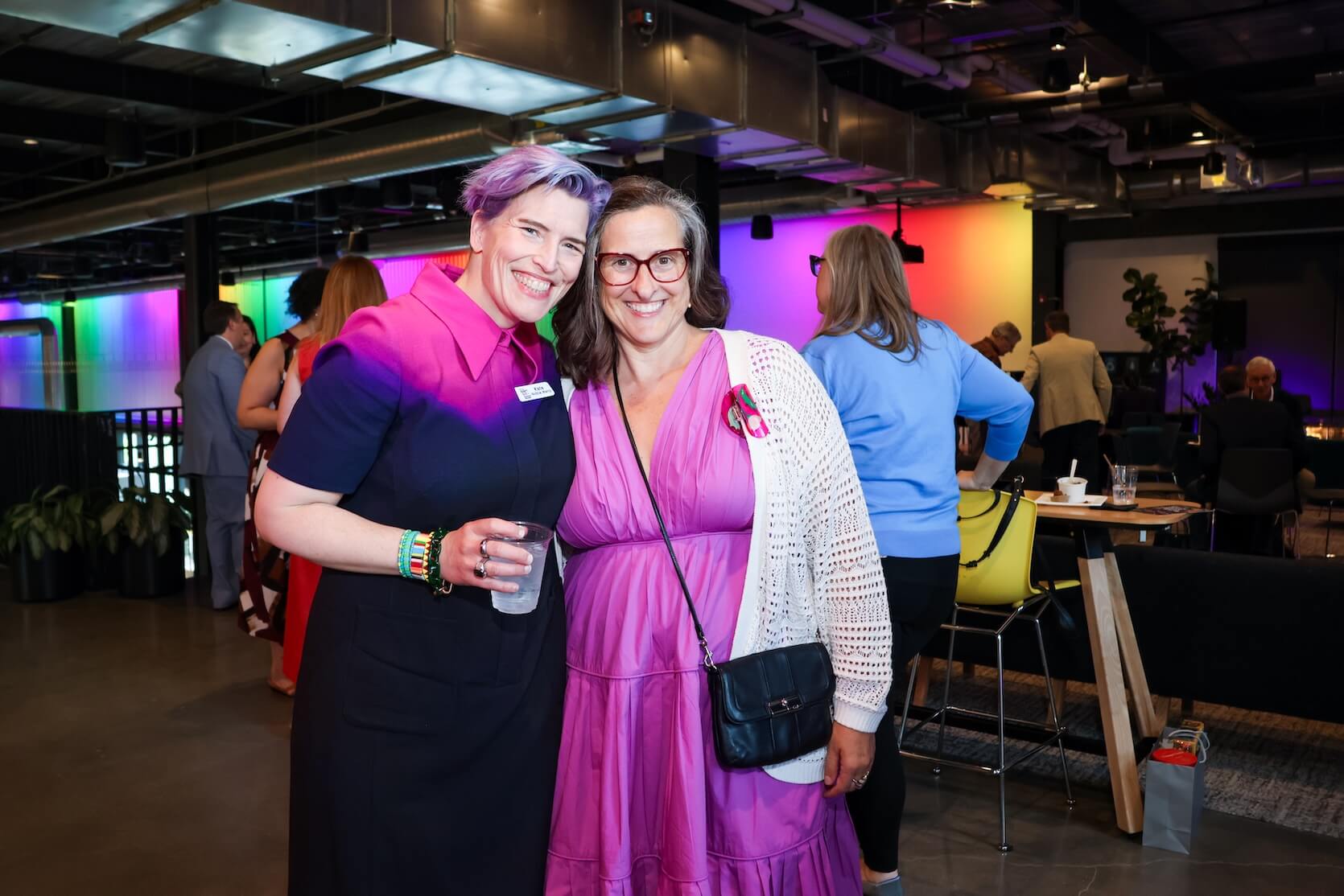 Two white women standing close together, smiling at the camera. Left has short purple hair, wearing a mostly black dress with small sections of pink and purple at the top. Right has long wavy brown and silver hair, wearing a long pink dress with a white cardigan over it.
