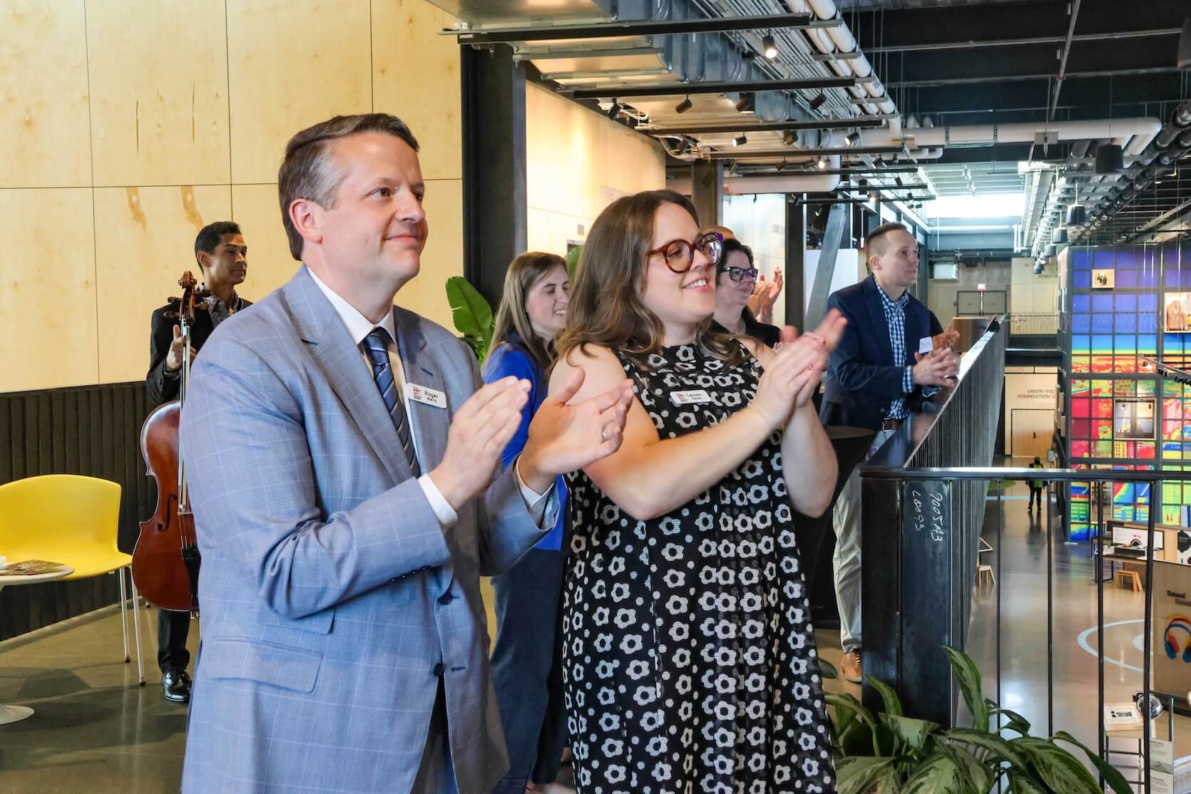 A white man and white woman standing beside each other, wearing a suit and dress, clapping after the speaker's remarks. More people stand behind them in the background.