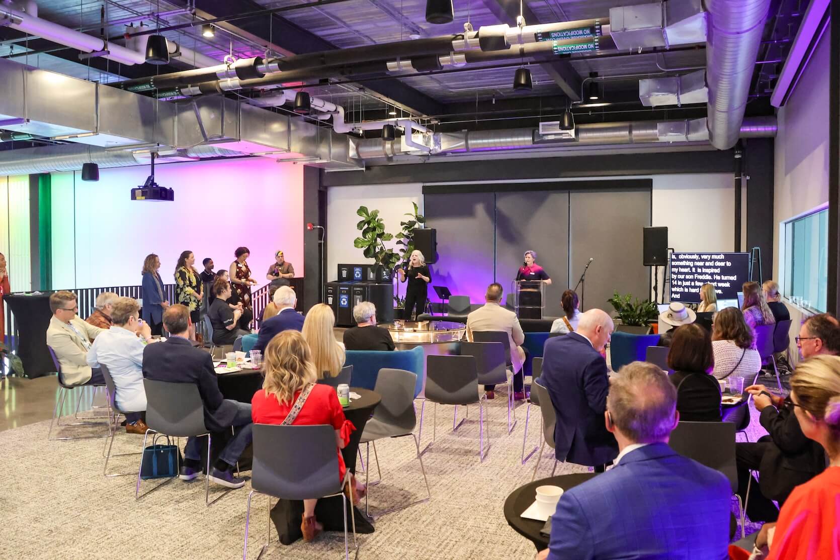 Back view of a large group of people sitting and standing, focusing on the speaker. There is also an ASL interpreter and Live Captions.