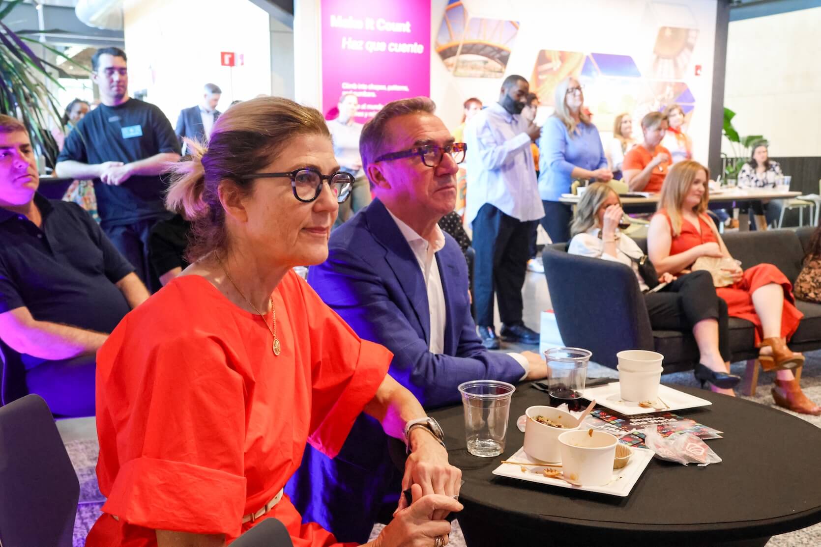 A white man and white woman sit at a table, focusing on the speaker. More people in the background.