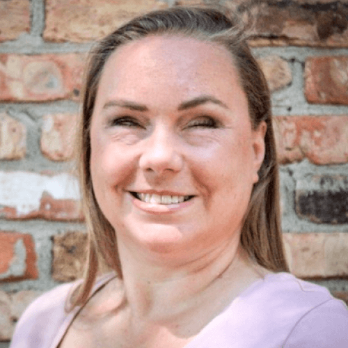 A white woman with straight blonde hair, standing outside in front of a brick wall, smiling. She is wearing a pink top.