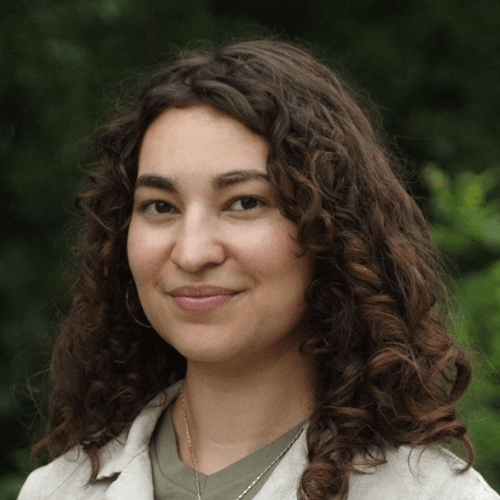 A Latina woman with long brown curly hair, wearing a green top and a jacket or some other top over it. She is standing outside in front of greenery.