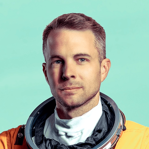 A white man with short and slicked or brushed back brown and gray hair, wearing an orange astronaut suit and holding the helmet between his arm and side. He is standing outside in what looks like an open field with some houses and other buildings, and trees and mountains very far away behind him.