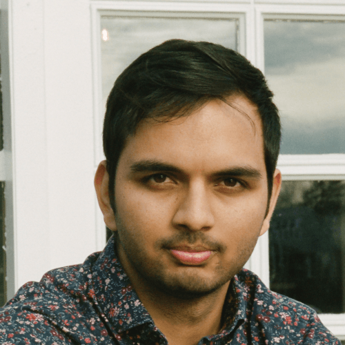 Rohan is a South-Asian man with short dark hair and a short, trimmed beard. He is wearing a heathered blue crewneck sweater with long sleeves folded up past the elbow, paired with grey straight-leg pants and white sneakers.