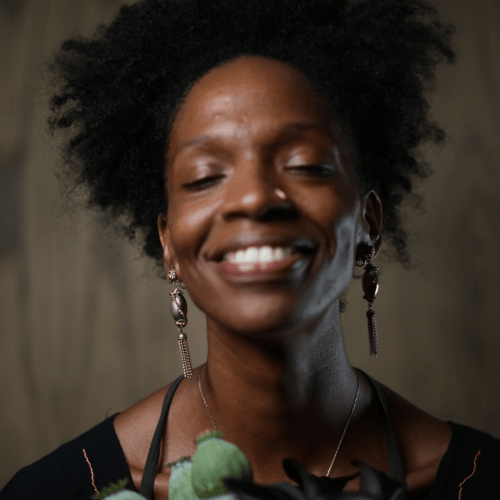 Courtney is a tall Black woman, standing six feet tall, with a curly afro that halos around her head. She is wearing a sleeveless periwinkle blue A-line dress with a mock turtleneck, a circular detail with a subtle sheen at one side of the waist, and a satin stripe running down the center. The dress falls just above her knees.