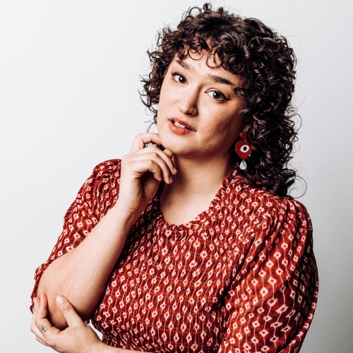 A Latinx person with curly brown hair, wearing black eyeliner, an orange lip, long dangling earrings, and a red-orange dress with a graphic print. They are standing in front of a gray wall, chin resting on their fingertips.