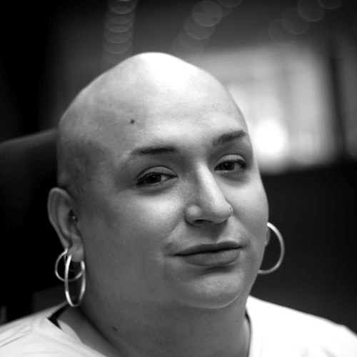 Grayscale photo. A bald Latina with two silver hoops in each ear, wearing a white shirt.