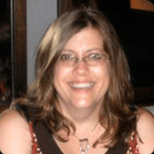 A white woman with shoulder length wavy brown hair, wearing glasses, a necklace, and a graphic tank top. She is smiling, sitting in a room.