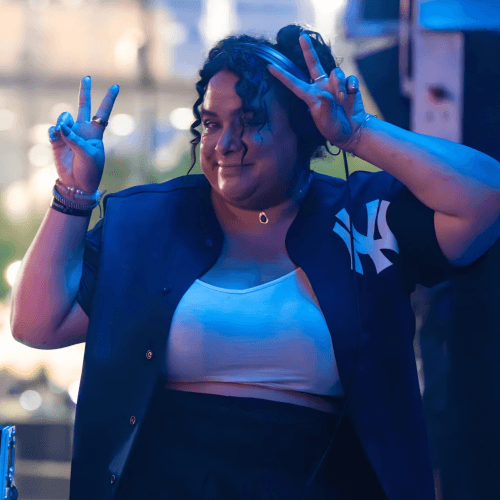 A woman standing outside by a DJ setup, her hair up, wearing an unbuttoned sports jersey of sorts, a white crop, and black bottoms, as well as a set of headphones. She is holding up two peace sign hand gestures.