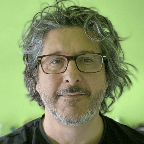 A white man with shaggy curly gray hair and a beard, wearing glasses and a black shirt. The background is green and there’s a bit of a green hue over the photo.
