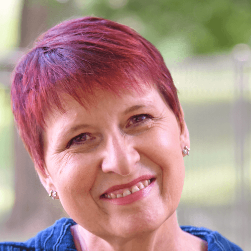 A white woman with pixie cut pink hair and pink lipstick, smiling. She is wearing a blue top. The photo is taken outside.