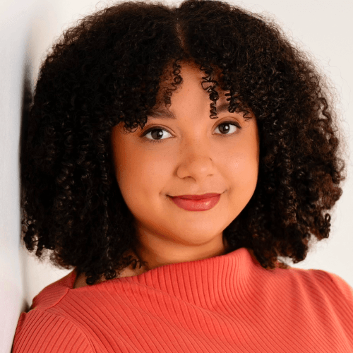 A light skinned woman with very curly black hair, minimal makeup, wearing a coral top.