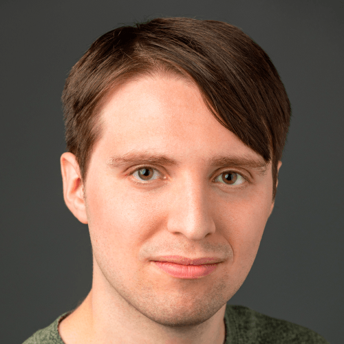 A white man with short brown hair, wearing a green top, in front of a dark wall.