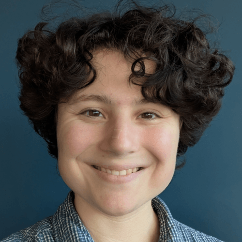 A white person with short curly black hair, wearing a blue and white gingham top, smiling in front of a dark gray-blue wall.
