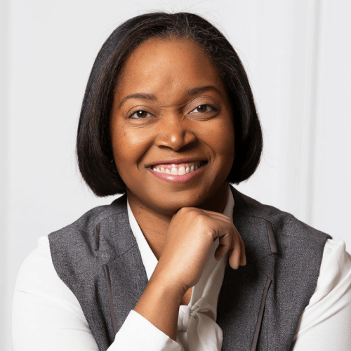 A Black woman with bob length straight black hair, wearing a long sleeve cream top with a gray vest or sleeveless jacket on top, smiling with her chin resting on the top of her hand. She is sitting in front of a white wall.